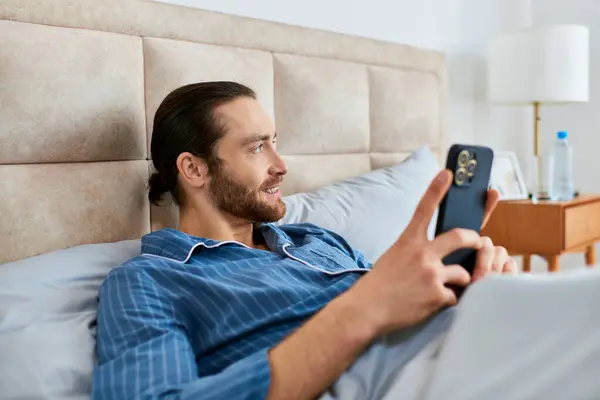Un hombre acostado en la cama, sosteniendo pacíficamente un teléfono celular mientras disfruta de un momento de relajación en la mañana. - foto de stock