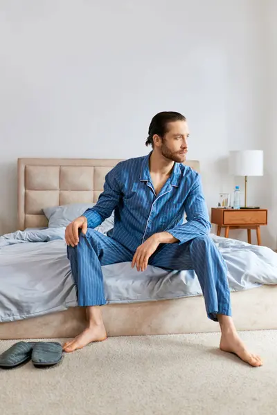 Un hombre guapo en pijama se sienta serenamente encima de una cama. - foto de stock