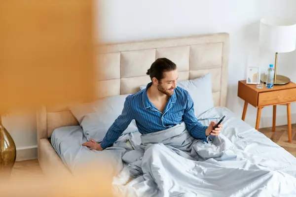 Un uomo si siede sul suo letto, assorto nello schermo del suo cellulare. — Foto stock