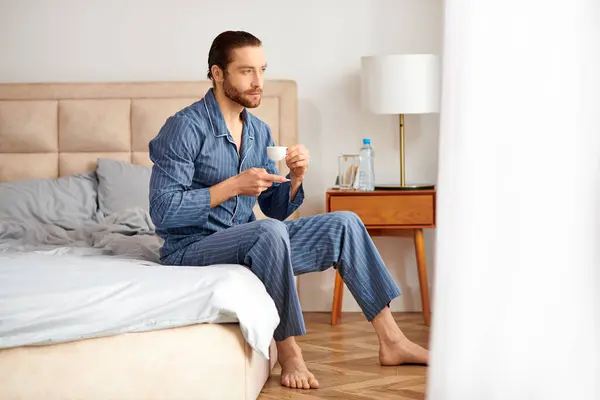 Um homem sentado em uma cama, desfrutando pacificamente de uma xícara de café em um ambiente acolhedor da manhã. — Fotografia de Stock