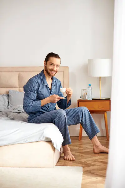 Un bell'uomo seduto su un letto, sorseggiando pacificamente una tazza di caffè. — Foto stock