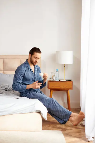 Handsome man peacefully sits on bed, sipping coffee in morning light. — Stock Photo
