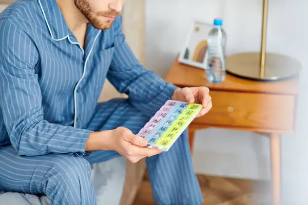 A man in pajamas sits on bed, deep in thought while holding a pill case. — Stock Photo