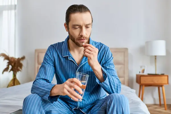 Um homem sentado em uma cama, bebendo pacificamente um copo de água com pílula. — Fotografia de Stock