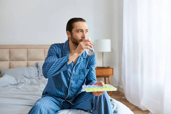 Bonito homem sentado na cama, bebendo água e pílulas. — Fotografia de Stock
