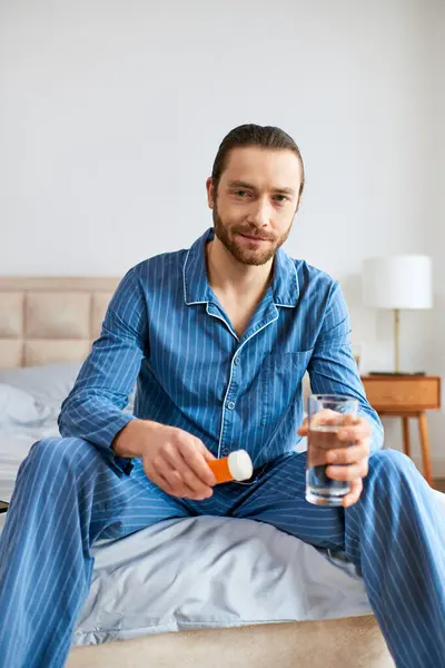 Bonito homem sentado na cama, segurando pacificamente vidro de água e pílulas. — Fotografia de Stock