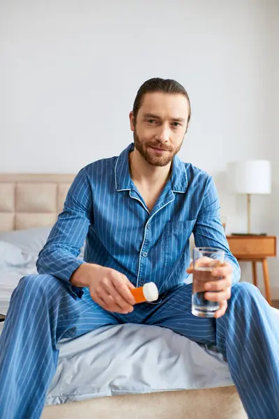 Hombre guapo sentado en la cama, sosteniendo pacíficamente un vaso de agua y pastillas. — Stock Photo