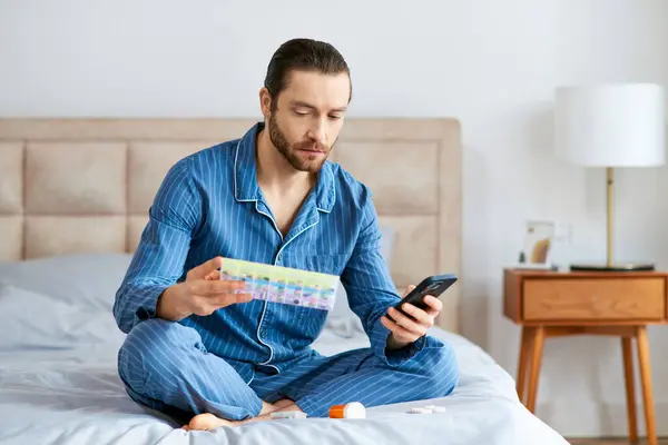 Bonito homem em casa sentado na cama com atenção fixa no celular e caixa de pílula. — Fotografia de Stock