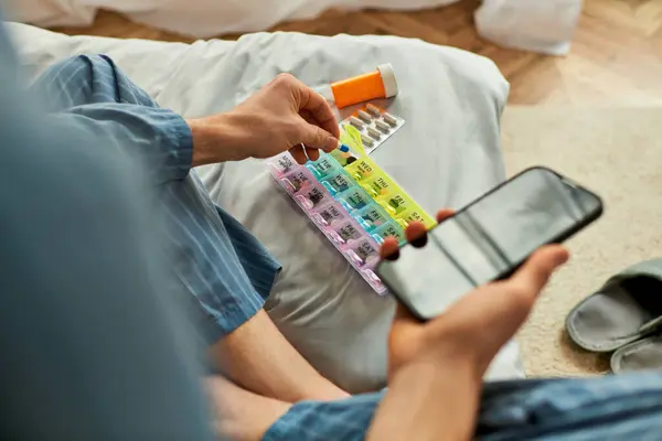 Un hombre sentado con su teléfono y pastillas en un sofá acogedor. - foto de stock