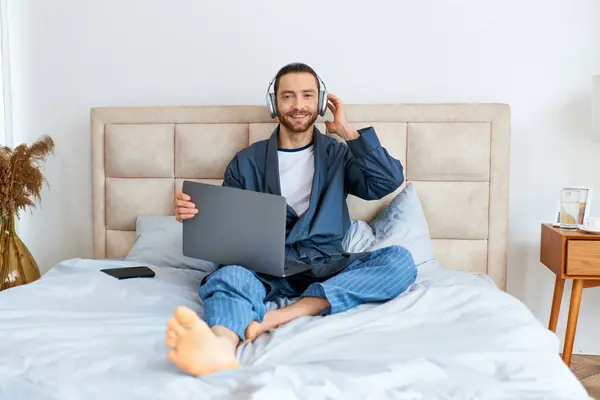 A man sitting on a bed, using a laptop and wearing headphones. — Stock Photo