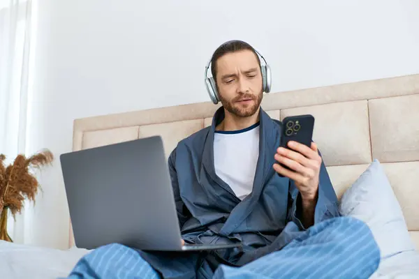 A man enjoys the morning, using a laptop and headphones on a bed. — Stock Photo