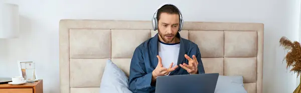 Un hombre sentado en una cama, profundamente centrado en el uso de un ordenador portátil. - foto de stock