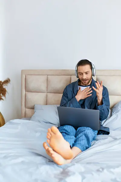 Un hombre sentado en una cama, absorto en el uso de una computadora portátil. - foto de stock