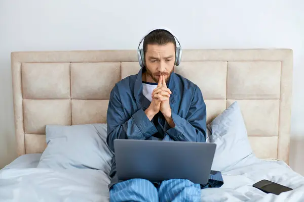 Un homme assis sur un lit, portant un casque, utilisant un ordinateur portable. — Photo de stock