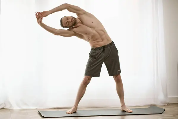 Hombre guapo parado en la esterilla de yoga, practicando yoga frente a la ventana. - foto de stock