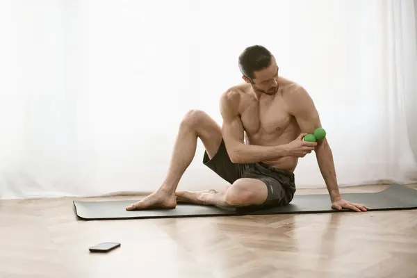 Homem sem camisa senta-se no tapete de ioga, segurando uma bola de massagem verde. — Fotografia de Stock