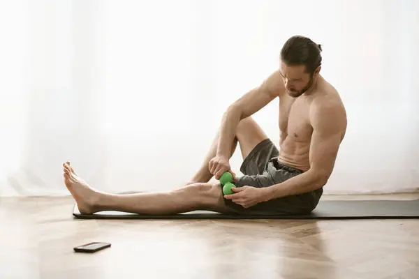 Un hombre se sienta en el suelo, sosteniendo una bola de masaje en su mano, practicando yoga en casa. - foto de stock