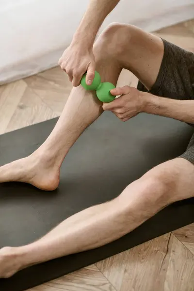 Un hombre se sienta en una esterilla sosteniendo una bola de masaje verde mientras practica yoga en la comodidad de su hogar. - foto de stock