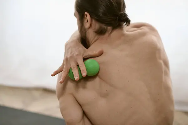 Un hombre con una bola de masaje verde en la mano derecha, haciendo yoga en casa. - foto de stock
