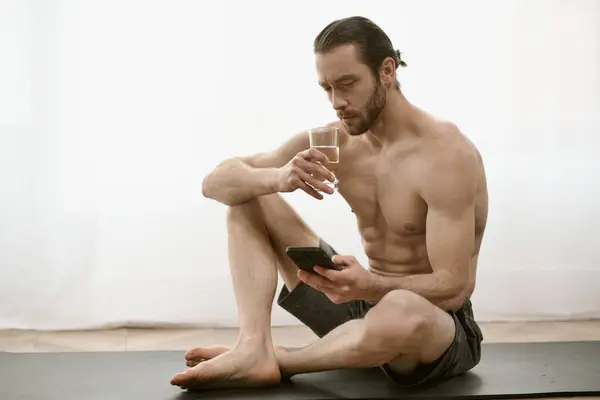 Shirtless man sitting on yoga mat, staring at cell phone. — Stock Photo