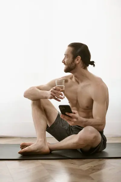 Un hombre se sienta en una esterilla de yoga, sosteniendo pacíficamente un teléfono celular. - foto de stock