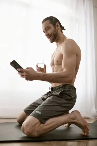 Un hombre sin camisa sentado en una esterilla de yoga, sosteniendo un teléfono celular, dedicado a la meditación matutina. - foto de stock