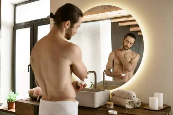 Un hombre se para frente a un espejo del baño, aplicando productos para el cuidado de la piel. - foto de stock