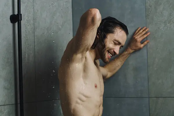 Un hombre sin camisa tomando una ducha refrescante en un baño en casa. - foto de stock