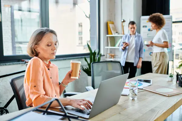 Woman enjoying coffee while working on laptop, with her diverse colleagues on backdrop. — Stock Photo