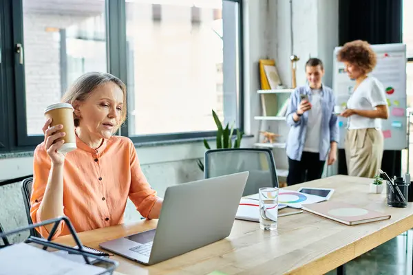 A woman fully engrossed in her work, sits before a laptop in a modern office space, with her diverse colleagues on backdrop. — Stock Photo