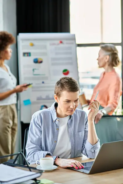 Short haired businesswoman engrossed in work on a laptop computer, with her diverse colleagues on backdrop. — Stock Photo
