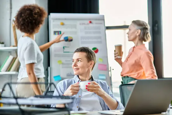 Devoted businesswoman engrossed in work on a laptop computer, with her diverse colleagues on backdrop. — Stock Photo