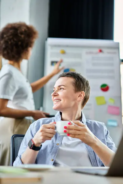 Attractive businesswoman engrossed in work on a laptop computer, with her african american colleague on backdrop. — Stock Photo