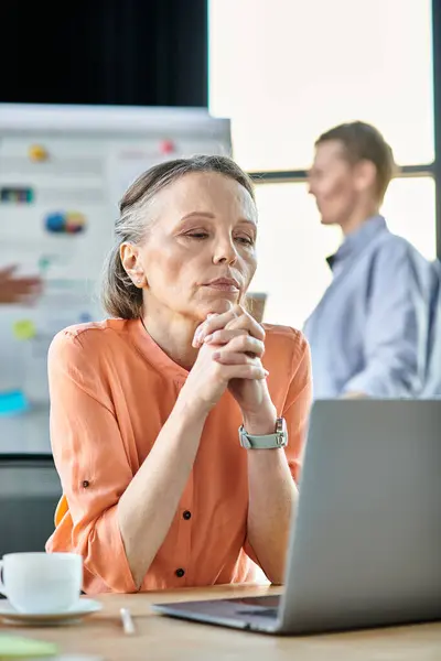 Une femme concentrée sur son ordinateur portable, immergée dans le travail dans un cadre de bureau moderne, avec sa collègue en toile de fond. — Photo de stock