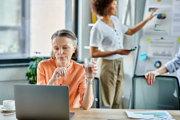 Mature woman focused on her laptop, immersed in work in a modern office setting, with her african american colleague on backdrop. — Stock Photo