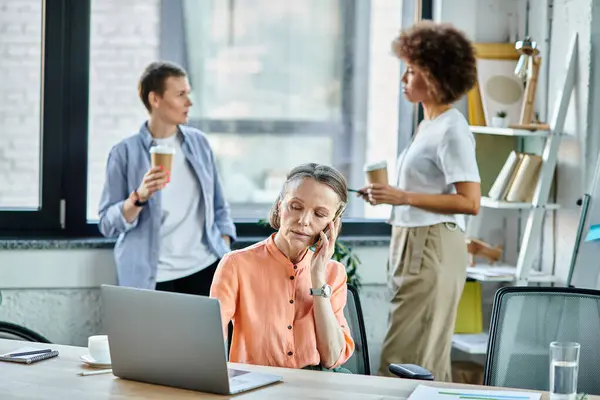 Mature businesswoman talking on phone while using laptop, with her diverse colleagues on backdrop. — Stock Photo