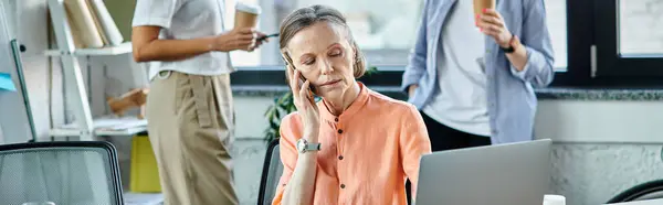 Dedicated businesswoman talking on phone while using laptop, with her diverse colleagues on backdrop. — Stock Photo