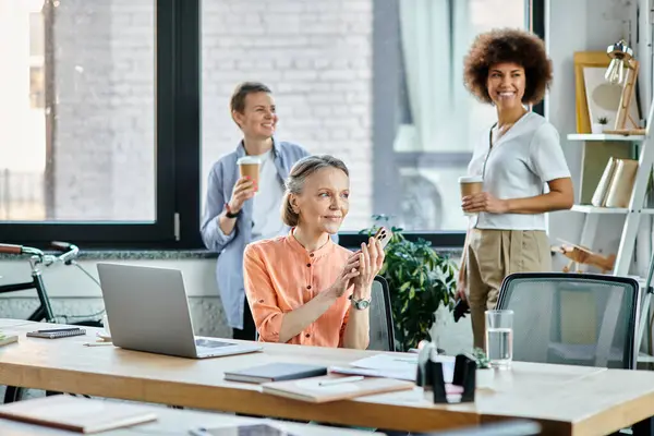 Jolly businesswoman talking on phone while using laptop, with her diverse colleagues on backdrop. — Stock Photo