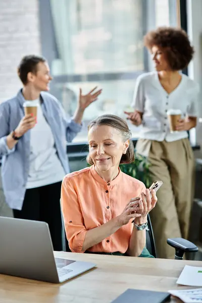 Merry businesswoman talking on phone while using laptop, with her diverse colleagues on backdrop. — Stock Photo