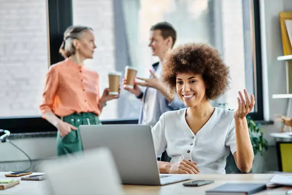 Joyous african american businesswoman using laptop, with her diverse colleagues on backdrop. — Stock Photo