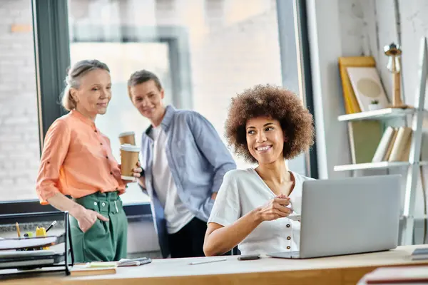 Attraente donna d'affari afroamericana che utilizza il computer portatile, con i suoi diversi colleghi sullo sfondo. — Foto stock