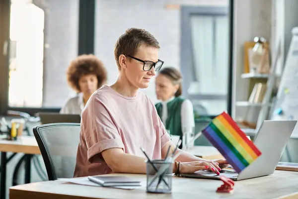 Appealing woman engrossed in work, with a laptop in front of her, with her diverse colleagues on backdrop, pride flag. — Stock Photo