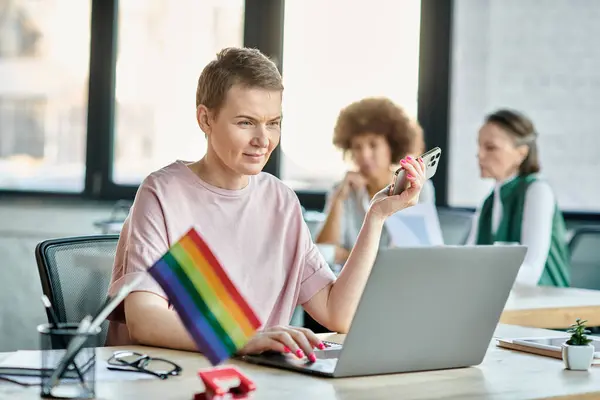 Mujer bonita absorta en el trabajo, con una computadora portátil frente a ella, con sus diversos colegas en el fondo, la bandera del orgullo. - foto de stock