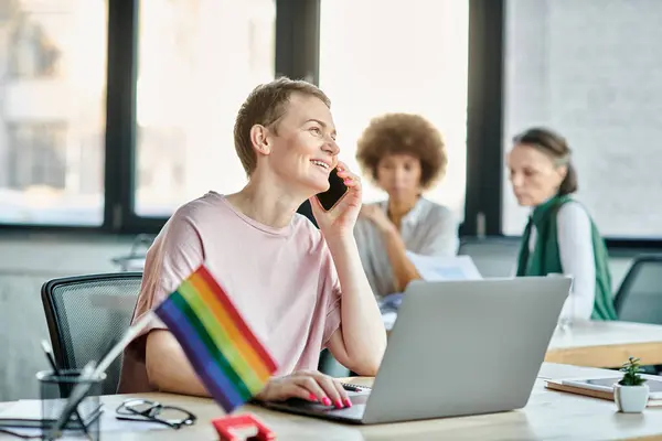 Jolly woman engrossed in work, with a laptop in front of her, with her diverse colleagues on backdrop, pride flag. — Stock Photo