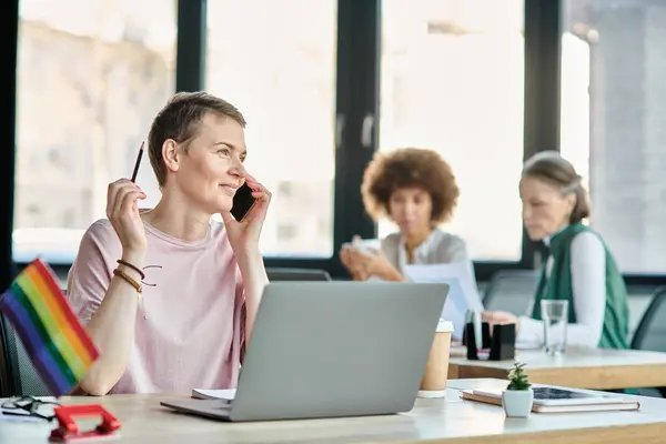 Cheerful woman engrossed in work, with a laptop in front of her, with her diverse colleagues on backdrop, pride flag. — Stock Photo