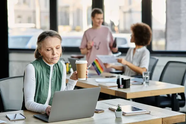 Beautiful mature woman works on a laptop at a table, with her diverse colleagues on backdrop. — Stock Photo
