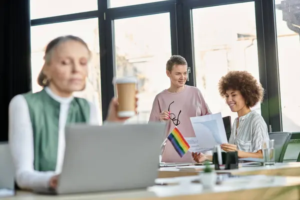 Blurred mature woman works on a laptop at a table, with her focused diverse colleagues on backdrop. — Stock Photo