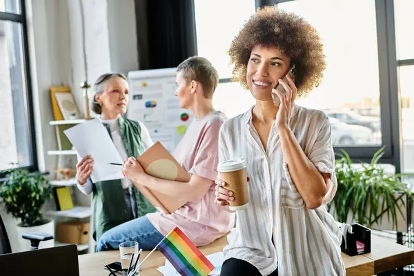 African american woman juggles a cell phone conversation and a cup of coffee, with her diverse colleagues. — Stock Photo