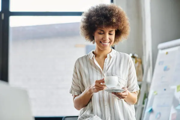 Mujer afroamericana, elegante y enfocada, se relaja con una taza de café humeante en la mano. - foto de stock