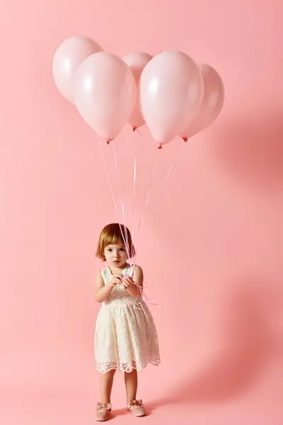 Adorable girl in white dress holding pink balloons on a pink background. — Stock Photo